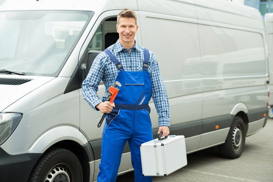 An Absolute Plumbing technician in blue overalls holding a pipe wrench in front of a service van